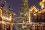 A festive scene featuring a large Christmas tree adorned with lights and ornaments, surrounded by snow-covered wooden houses illuminated with string lights. A wooden walkway connects the buildings, adding to the cozy, wintry atmosphere.