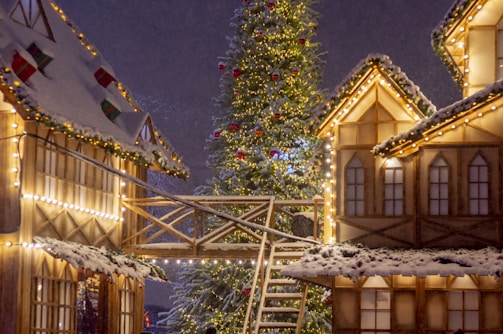 A festive scene featuring a large Christmas tree adorned with lights and ornaments, surrounded by snow-covered wooden houses illuminated with string lights. A wooden walkway connects the buildings, adding to the cozy, wintry atmosphere.