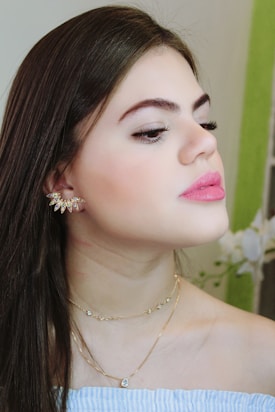 A person with long brown hair is wearing delicate gold jewelry, including earrings and a necklace. The expression is serene and contemplative, with eyes gently closed and lips slightly parted. Light blue and white striped clothing is visible, along with a blurred background containing a green element and some flowers.