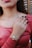 A close-up of a person wearing multiple elegant gold rings and bracelets, including a distinctive bracelet with an eye symbol. The person has painted black nails and is dressed in a maroon top. The background includes what appears to be wooden shutters or a textured surface.