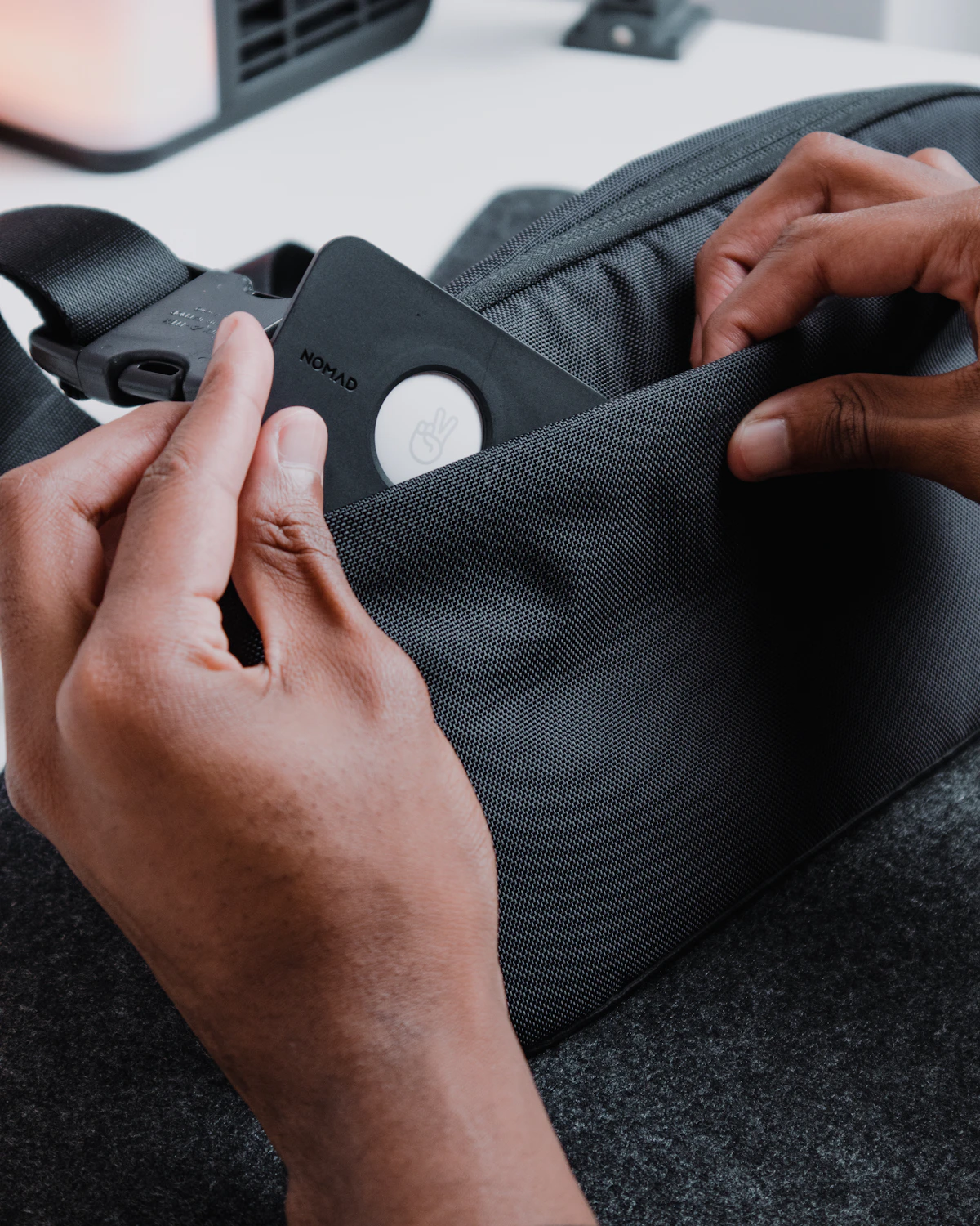 Hands placing an Apple AirTag inside the pocket of a grey backpack before a trip