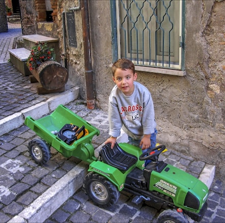A young child is sitting on a toy green tractor with a trailer attached, parked on a cobblestone street in front of an old stone building. The child is wearing a grey sweatshirt with red lettering. Nearby, there is a large log with flowers in a planter.