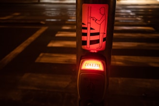 A pedestrian crossing button at night with an illuminated sign displaying instructions in Czech. The surroundings are dimly lit and the crosswalk is visible in the background.