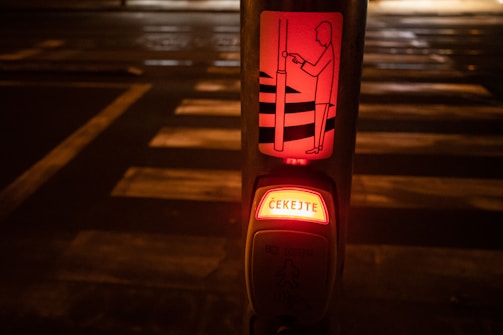 A pedestrian crossing button at night with an illuminated sign displaying instructions in Czech. The surroundings are dimly lit and the crosswalk is visible in the background.