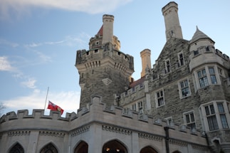 A historic stone castle with intricate architectural details and tall towers stands against a clear blue sky. A flag flies at half-mast on a pole in front of the castle.