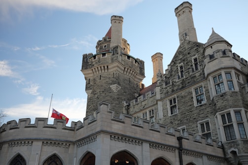 A historic stone castle with intricate architectural details and tall towers stands against a clear blue sky. A flag flies at half-mast on a pole in front of the castle.