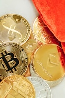 Several physical representations of cryptocurrencies such as Bitcoin, Ethereum, and others are scattered on a surface. The coins are predominantly golden, with a few silver-colored ones, and are partially covered by a vibrant orange fabric.