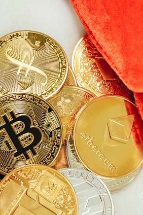 Several physical representations of cryptocurrencies such as Bitcoin, Ethereum, and others are scattered on a surface. The coins are predominantly golden, with a few silver-colored ones, and are partially covered by a vibrant orange fabric.