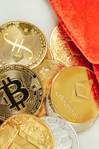 Several physical representations of cryptocurrencies such as Bitcoin, Ethereum, and others are scattered on a surface. The coins are predominantly golden, with a few silver-colored ones, and are partially covered by a vibrant orange fabric.