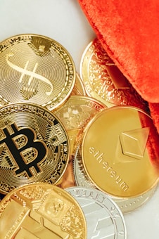 Several physical representations of cryptocurrencies such as Bitcoin, Ethereum, and others are scattered on a surface. The coins are predominantly golden, with a few silver-colored ones, and are partially covered by a vibrant orange fabric.