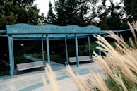 A pathway leads through a park area surrounded by lush greenery and tall trees. A structure spans over the path with a sign that reads 'THE JOURNEY IS THE REWARD.' Long grasses sway in the foreground, adding a natural, serene element to the scene.