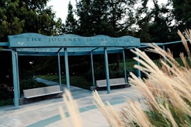 A pathway leads through a park area surrounded by lush greenery and tall trees. A structure spans over the path with a sign that reads 'THE JOURNEY IS THE REWARD.' Long grasses sway in the foreground, adding a natural, serene element to the scene.