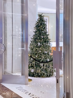 A beautifully decorated Christmas tree adorned with white lights and ornaments stands elegantly in a polished, luxurious setting. The surrounding area is pristine, featuring marble flooring with the name 'Tiffany & Co.' visible, suggesting a high-end retail environment.