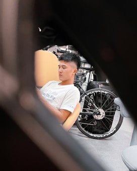 A person is sitting in a relaxed posture on a beige chair. A bicycle with a large wheel featuring the word 'SCULTURA' is positioned beside them. The image appears to be taken through a dark foreground, creating a framed effect.