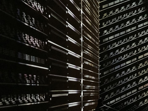 A dark room filled with rows of server racks illuminated by tiny lights and reflections. The metallic surfaces create a sense of symmetry and order, while the low light adds a mysterious ambiance.