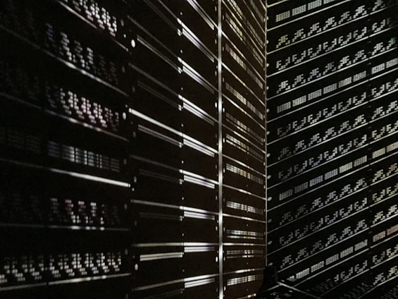 A dark room filled with rows of server racks illuminated by tiny lights and reflections. The metallic surfaces create a sense of symmetry and order, while the low light adds a mysterious ambiance.