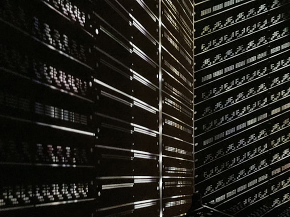 A dark room filled with rows of server racks illuminated by tiny lights and reflections. The metallic surfaces create a sense of symmetry and order, while the low light adds a mysterious ambiance.