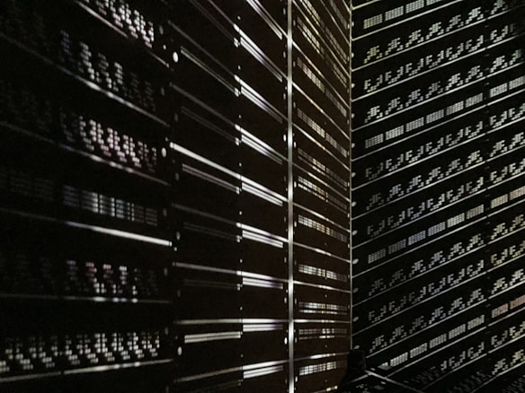 A dark room filled with rows of server racks illuminated by tiny lights and reflections. The metallic surfaces create a sense of symmetry and order, while the low light adds a mysterious ambiance.