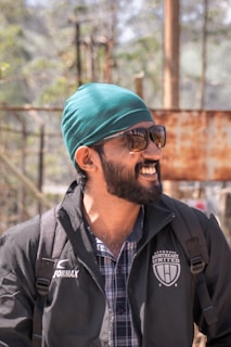 A person with a beard and sunglasses is wearing a turquoise headscarf and a jacket with a logo that reads 'Northeast United FC'. The individual appears to be outside, under natural light, with a blurred background of trees and structures.