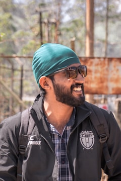 A person with a beard and sunglasses is wearing a turquoise headscarf and a jacket with a logo that reads 'Northeast United FC'. The individual appears to be outside, under natural light, with a blurred background of trees and structures.