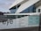 The image depicts a modern architectural building with a geometric design and large glass windows. The foreground shows signage with the word 'eye' and a list of facilities available, such as museum, film, shop, and restaurant. The background includes a waterfront view with additional buildings under a cloudy sky.