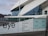 The image depicts a modern architectural building with a geometric design and large glass windows. The foreground shows signage with the word 'eye' and a list of facilities available, such as museum, film, shop, and restaurant. The background includes a waterfront view with additional buildings under a cloudy sky.