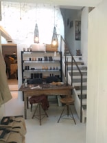 A cozy interior of a home or studio with a rustic wooden table and stools in the foreground. Shelves filled with various pottery items and decorative objects are set against a white textured wall. Two pendant lights hang from the ceiling, casting a warm, ambient glow. A staircase leads to an upper level, and natural light fills the space, enhancing the cozy and intimate atmosphere.