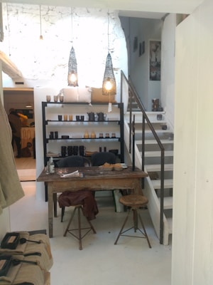 A cozy interior of a home or studio with a rustic wooden table and stools in the foreground. Shelves filled with various pottery items and decorative objects are set against a white textured wall. Two pendant lights hang from the ceiling, casting a warm, ambient glow. A staircase leads to an upper level, and natural light fills the space, enhancing the cozy and intimate atmosphere.