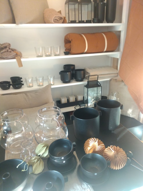 A collection of home decor items and cookware is displayed on shelves and a table. The shelves contain folded blankets, glass candle holders, and decorative jars. The table features black pots, bowls, and paper decorations. The overall arrangement suggests a minimalistic aesthetic with earthy tones.