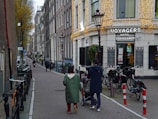 A street scene features a couple walking with a stroller past a building decorated with festive lights. Bikes are parked along the sidewalk, and various people are seen further down the street. The building on the right displays signs for a hotel and coffeeshop named 'Voyagers'. Autumn leaves are visible on the trees lining the street.