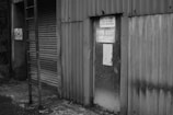 An industrial building features a corrugated metal wall with a fire exit door. Signs warn of required eye protection and emphasize keeping the fire exit clear. A ladder is attached to the wall, and there is a warning sign indicating danger.