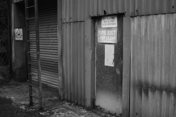 An industrial building features a corrugated metal wall with a fire exit door. Signs warn of required eye protection and emphasize keeping the fire exit clear. A ladder is attached to the wall, and there is a warning sign indicating danger.
