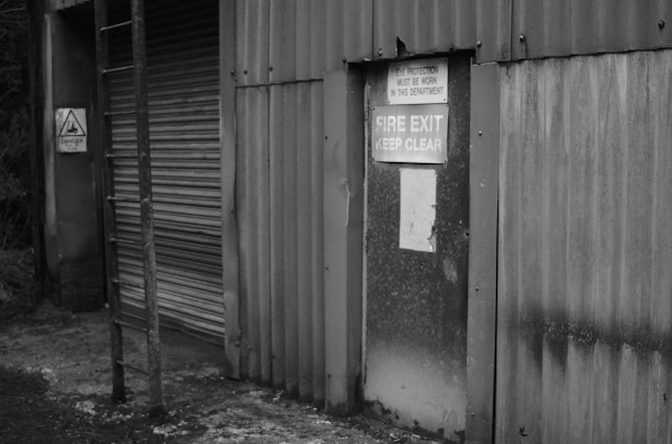 An industrial building features a corrugated metal wall with a fire exit door. Signs warn of required eye protection and emphasize keeping the fire exit clear. A ladder is attached to the wall, and there is a warning sign indicating danger.