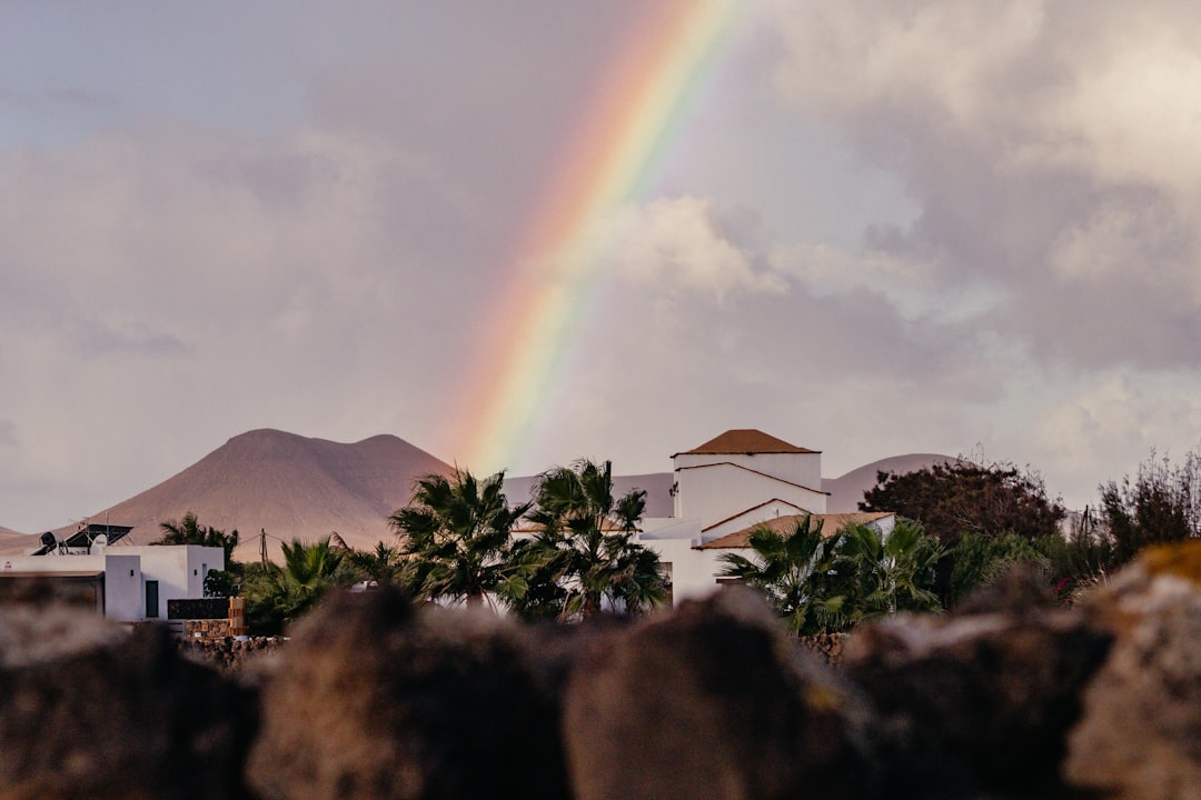 a rainbow in the sky over a building and palm trees, rainbow over lajares and the hills of fuerteventura