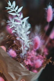 A bouquet of dried flowers featuring delicate, fluffy pink blooms and muted brown and white leaves. The composition is wrapped in brown paper, creating a rustic and elegant appearance.