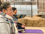 A person wearing glasses and a colorful scarf is sitting with eyes closed, appearing to meditate on a purple yoga mat. The background features another person in a similar pose, with bales of hay and a greenhouse structure visible.