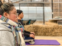 A person wearing glasses and a colorful scarf is sitting with eyes closed, appearing to meditate on a purple yoga mat. The background features another person in a similar pose, with bales of hay and a greenhouse structure visible.