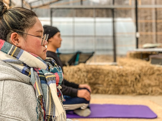 A person wearing glasses and a colorful scarf is sitting with eyes closed, appearing to meditate on a purple yoga mat. The background features another person in a similar pose, with bales of hay and a greenhouse structure visible.