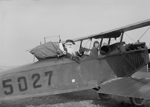 A vintage biplane with the registration number 5027 is on the ground. A person dressed as Santa Claus is sitting in the back seat with a large sack, appearing cheerful and playful. Another person, possibly a pilot, is sitting in the front seat wearing an aviator cap and goggles.