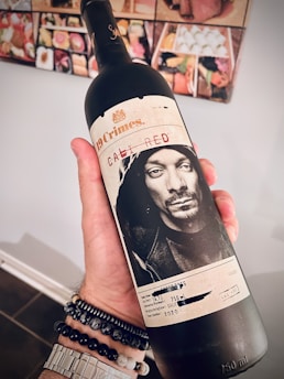 A hand is holding a wine bottle labeled 19 Crimes Cali Red, featuring a monochrome portrait of a person on the label. The background displays a wall with colorful square-shaped images, possibly food items or art. The hand is adorned with beaded bracelets and a watch.