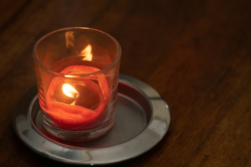 A lit candle nestled in a clear glass holder casts a warm glow. The candle sits on a metallic dish placed on a wooden surface, enhancing the cozy ambiance.