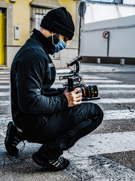 A person kneels on a crosswalk while handling a professional video camera. They are wearing dark clothing, a beanie, and a face mask. The background shows a building corner with a muted yellow wall and a small signpost.