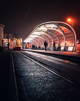 A modern tram station at night, illuminated with bright lights under a curved metal roof. A tram is parked on the tracks, and a few people are present, including one person using a wheelchair. The setting is urban with a warm, slightly foggy ambiance and reflections on the wet pavement.