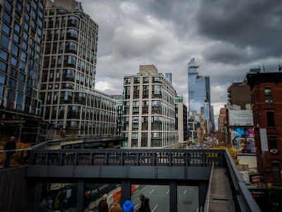 Urban landscape with tall, modern buildings under a cloudy sky. The scene is viewed from an elevated perspective, possibly a bridge or walkway, and features a mixture of architectural styles. People can be seen below and modern high-rises are visible in the distance.