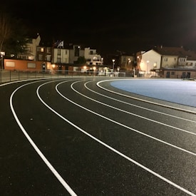 A curved running track is brightly lit at night. The black asphalt surface features white lane markings curving to the right. Adjacent to the track is a blue area, possibly another sports field. In the background are buildings with warm lights visible in windows, suggesting a residential area.