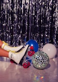 A pair of retro white roller skates with yellow laces and red wheels rests on a shiny floor. Next to them, a mosaic disco ball reflects light, and there are blue and white balloons scattered around. The background features a shimmering silver curtain, creating a festive or party-like atmosphere.