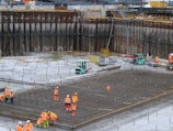 A construction site with several workers wearing orange safety vests and helmets. They are working on a large framework of rebar for concrete reinforcement. In the background, there are forklifts and construction materials scattered around. The site is surrounded by high retaining walls indicating excavation work.