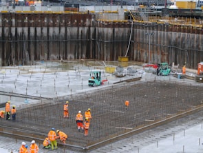A construction site with several workers wearing orange safety vests and helmets. They are working on a large framework of rebar for concrete reinforcement. In the background, there are forklifts and construction materials scattered around. The site is surrounded by high retaining walls indicating excavation work.