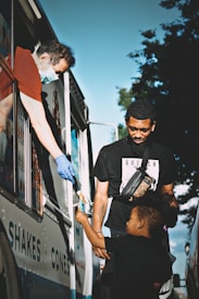 A child reaches out to receive an ice cream from a vendor in an ice cream truck, while an adult stands nearby, both appearing happy and engaged in the moment. The vendor wears a mask and gloves, indicating attention to hygiene and safety.