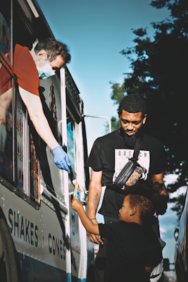 A child reaches out to receive an ice cream from a vendor in an ice cream truck, while an adult stands nearby, both appearing happy and engaged in the moment. The vendor wears a mask and gloves, indicating attention to hygiene and safety.